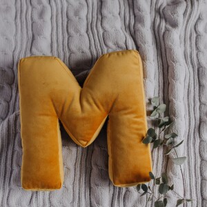 May include: A mustard yellow velvet throw pillow shaped like the capital letter M. The pillow has a soft, plush texture and is positioned next to a thin, bare branch with small, round, muted green leaves, resting on a light gray cable knit fabric.