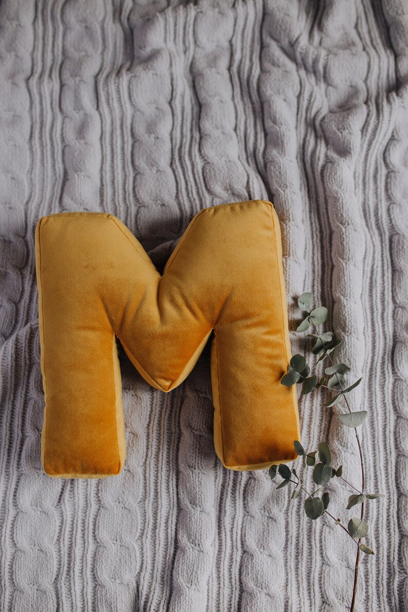 May include: A mustard yellow velvet throw pillow shaped like the capital letter M. The pillow has a soft, plush texture and is positioned next to a thin, bare branch with small, round, muted green leaves, resting on a light gray cable knit fabric.