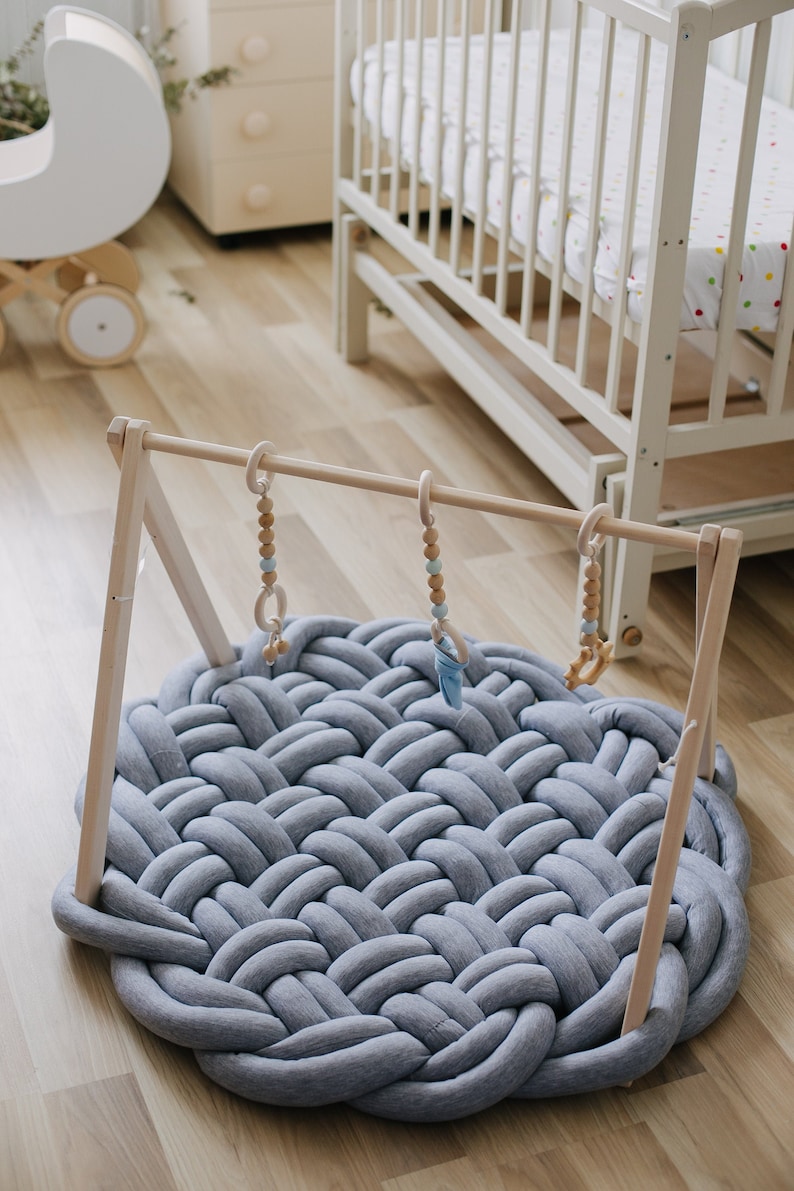 May include: A gray, round baby play mat with a braided design. A wooden baby gym with hanging toys is positioned above the mat. The toys include wooden rings and beads. A white crib is in the background.