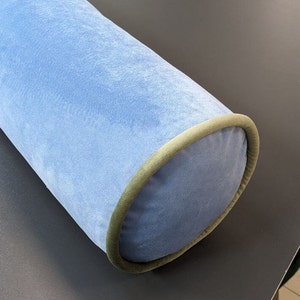 May include: A light blue cylindrical bolster pillow with a soft, velvety texture. The ends are a darker blue with a green trim. This decorative pillow is designed for comfort and support, suitable for various home decor styles.