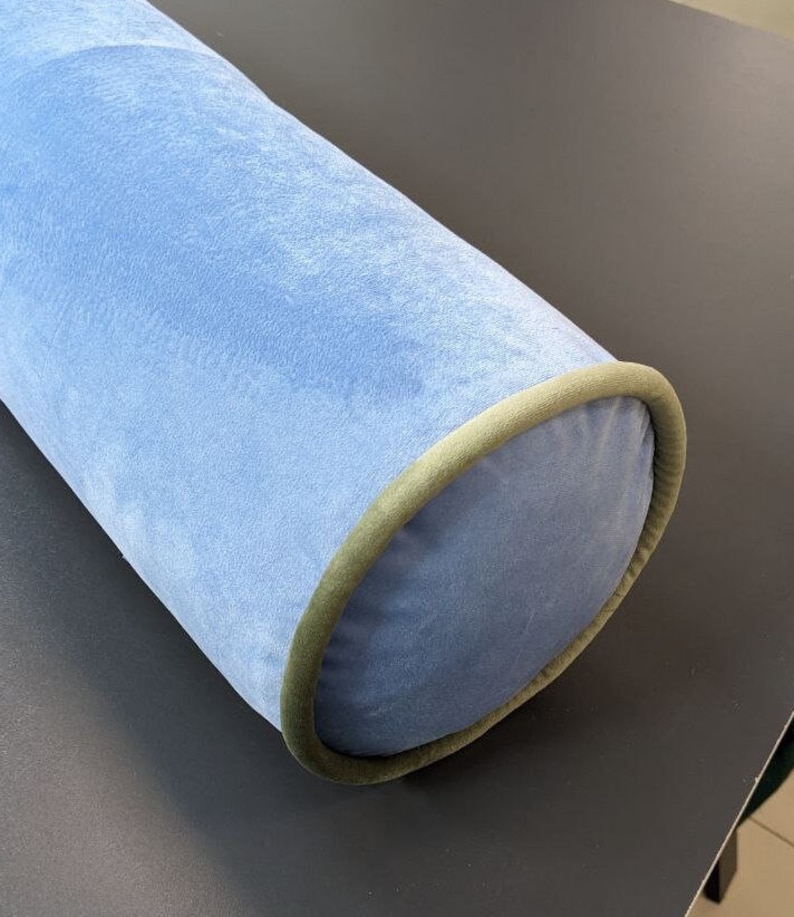 May include: A light blue cylindrical bolster pillow with a soft, velvety texture. The ends are a darker blue with a green trim. This decorative pillow is designed for comfort and support, suitable for various home decor styles.