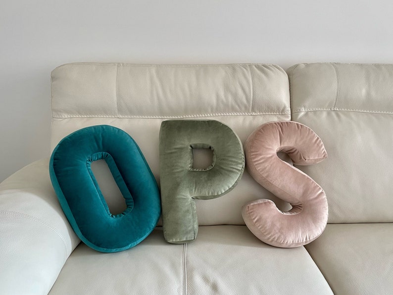 May include: Three plush letter-shaped pillows spelling the word OPS. The letter O is teal, the letter P is sage green, and the letter S is light pink. Each pillow is crafted from a soft velvet fabric with a smooth, rounded, and stuffed construction.