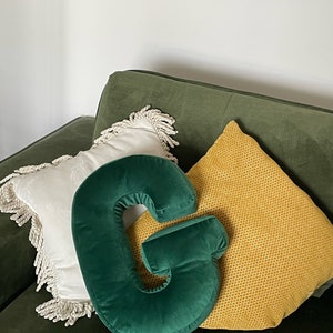 May include: A dark green velvet throw pillow shaped like the letter G, positioned in front of a white square pillow with fringe trim and a yellow textured square pillow, all resting on a dark green upholstered surface.