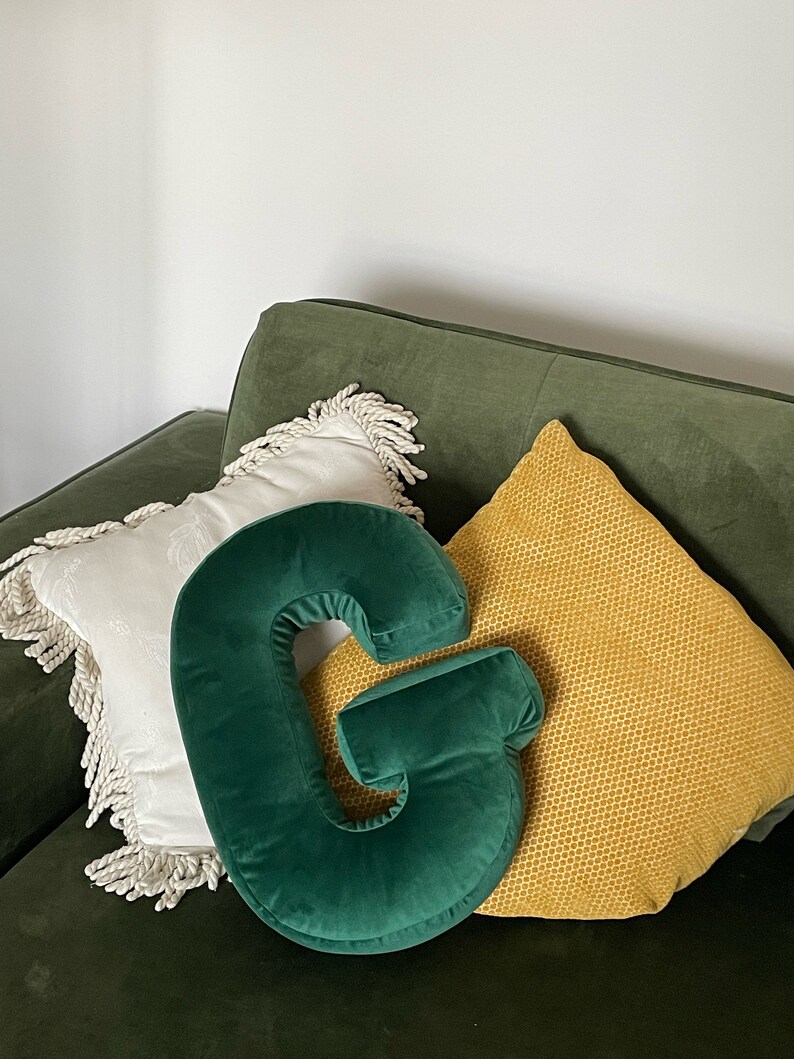 May include: A dark green velvet throw pillow shaped like the letter G, positioned in front of a white square pillow with fringe trim and a yellow textured square pillow, all resting on a dark green upholstered surface.