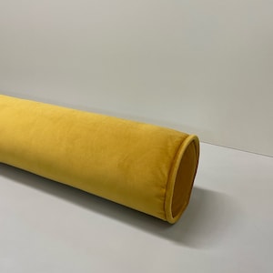 May include: A long, cylindrical bolster pillow in a vibrant mustard yellow. The pillow has a soft, velvety texture and a rounded end with a contrasting trim. The design is simple and elegant, suitable for home decor.