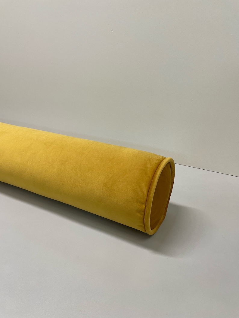 May include: A long, cylindrical bolster pillow in a vibrant mustard yellow. The pillow has a soft, velvety texture and a rounded end with a contrasting trim. The design is simple and elegant, suitable for home decor.