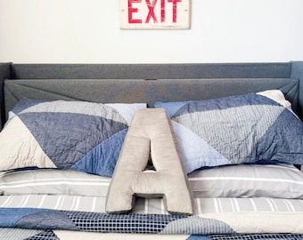 May include: A light gray plush letter A pillow sits on a bed with blue and white geometric patterned bedding. A dark gray upholstered headboard is behind the pillows, and a rectangular white sign with the red text EXIT is on the wall.