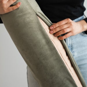 May include: Olive green velvet bolster pillow with a contrasting beige panel and visible stitching. The pillow is cylindrical and appears soft to the touch. The image shows a close-up of the pillow, held by a person.