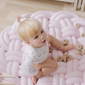 May include: A baby sits on a large, round, pink braided play mat. The baby is wearing a white onesie with a light blue pattern. Several wooden toy rings are scattered on the mat. The mat is on a light-colored wooden floor.