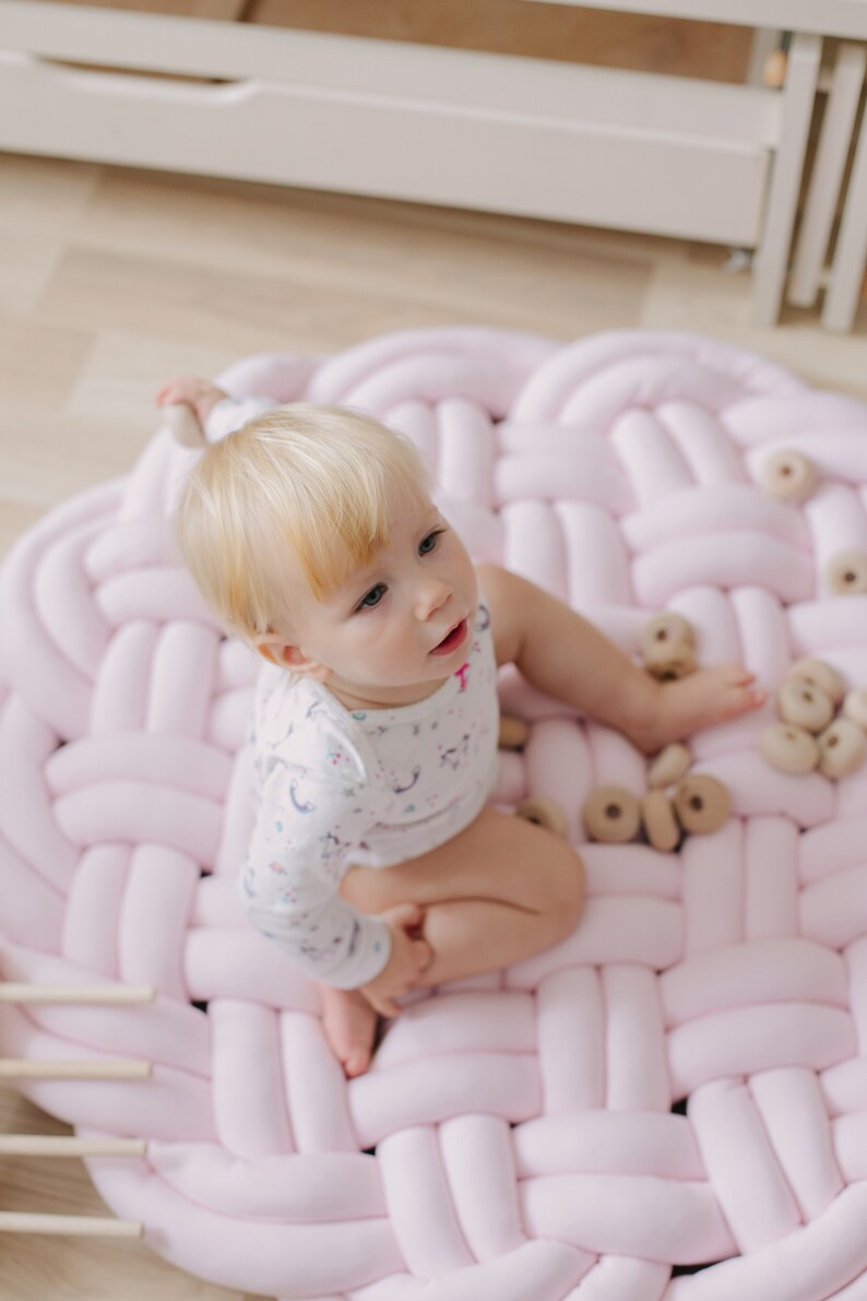 May include: A baby sits on a large, round, pink braided play mat. The baby is wearing a white onesie with a light blue pattern. Several wooden toy rings are scattered on the mat. The mat is on a light-colored wooden floor.