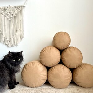 May include: A stack of six cylindrical brown throw pillows arranged in a pyramid shape. The pillows are made of a soft, plush fabric and have a subtle sheen.
