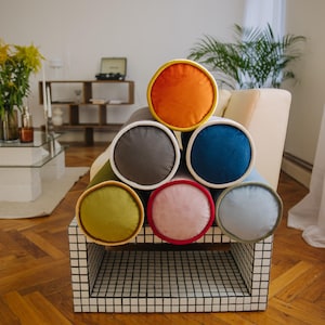 Könnte beinhalten: Ein Stapel aus sieben bunten, zylindrischen Kissen mit weißem Besatz, angeordnet in Pyramidenform auf einem Regal aus weißen und schwarzen Fliesen. Die Kissen sind aus Samt und in den Farben Orange, Grau, Blau, Grün, Rosa und Hellblau gehalten.