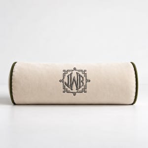 Könnte beinhalten: Ein zylindrisches Nackenkissen aus hellbeigem Samtstoff mit dunkelolivgrüner Paspelierung an den Kanten. In der Mitte befindet sich ein dunkelgraues gesticktes Monogramm mit den Buchstaben JWB in einem geometrischen quadratischen Rahmen.
