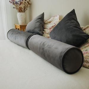 May include: A gray velvet bolster pillow and two matching triangular pillows rest on a white bedspread. The pillows are arranged on a bed with a floral patterned blanket and a vase of dried flowers in the background.