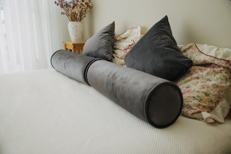 May include: A gray velvet bolster pillow and two matching triangular pillows rest on a white bedspread. The pillows are arranged on a bed with a floral patterned blanket and a vase of dried flowers in the background.