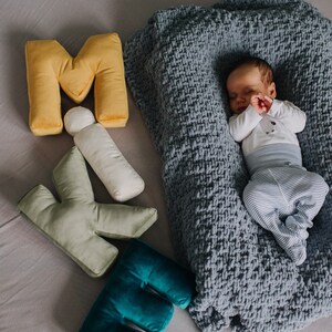 May include: A sleeping infant wearing a white long-sleeve shirt and blue and white striped pants, nestled in a grey chunky knit blanket, accompanied by plush letter-shaped pillows in yellow, sage green, and teal, plus a small white cylinder.
