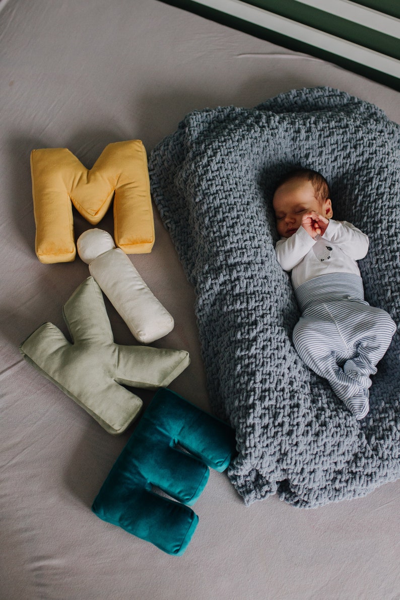 May include: A sleeping infant wearing a white long-sleeve shirt and blue and white striped pants, nestled in a grey chunky knit blanket, accompanied by plush letter-shaped pillows in yellow, sage green, and teal, plus a small white cylinder.