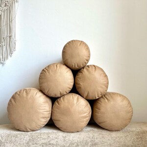 May include: A stack of six round, tan-colored throw pillows arranged in a pyramid shape. The pillows are made of a soft, plush fabric.