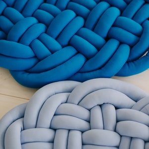 May include: Close-up of two braided round cushions. One is a vibrant blue, the other a soft, light blue. The cushions are made of a soft, plush material, with a woven design. Home decor item.