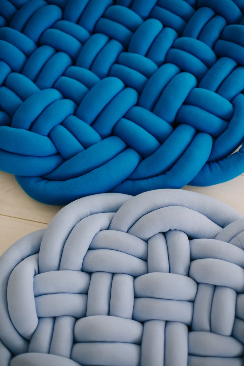 May include: Close-up of two braided round cushions. One is a vibrant blue, the other a soft, light blue. The cushions are made of a soft, plush material, with a woven design. Home decor item.