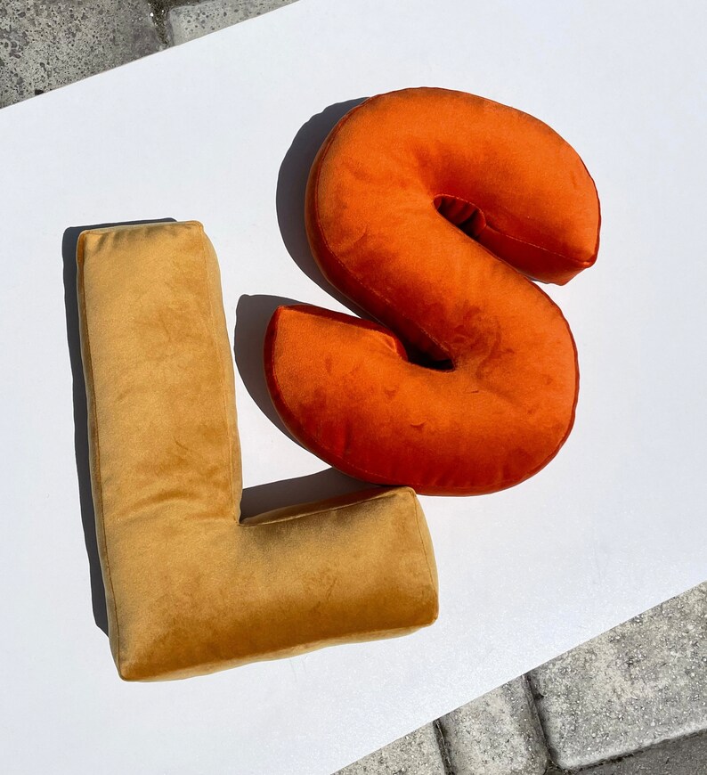 May include: A mustard yellow velvet letter L pillow positioned next to a burnt orange velvet letter S pillow. Both pillows feature a soft, plush texture and are shaped like the capital letters L and S, creating a decorative set of alphabet cushions.