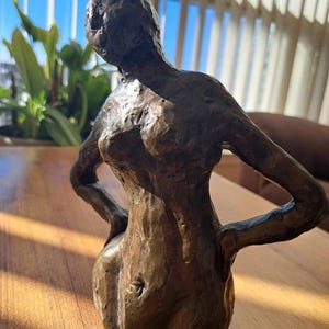 May include: A white ceramic mug with a black handle and the text "But First, Coffee" in a bold, black font. The mug is set against a blurred background. The mug appears to be approximately 10 cm tall.