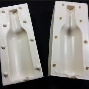 May include: Two white silicone molds for making bottle-shaped objects. Each mold has a smooth, hollow interior shaped like a bottle.