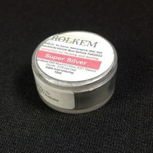 Könnte beinhalten: Ein kleines silbernes Glas mit einem weißen Etikett, auf dem "Super Silver" steht und das essbare Lebensmittelfarbe enthält. Das Etikett enthält auch die Zutaten, Gebrauchsanweisungen und ein Mindesthaltbarkeitsdatum von Februar 2022.