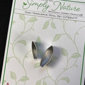 May include: Two silver daisy petal cutters on a white background with green vines. The packaging says "Simply Nature Botanically Correct Products Daisy Marguerite Petal Set CUT864-TT"