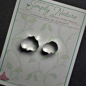 Könnte beinhalten: Zwei silberne Metall-Ausstecher in Form von Hortensienblütenblättern. Die Ausstecher befinden sich auf einem weißen Hintergrund mit einem grünen Rankenmuster. Der Text "Simply Nature" und "Hydrangea Petal Set CUT609-TT" ist auf den Hintergrund gedruckt.