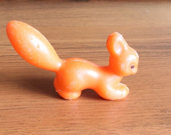 May include: An orange plastic squirrel figurine with a large, curved tail. The squirrel is in a side-facing pose, with its front paws slightly raised. The figurine has a small, dark eye and is set against a wooden surface.