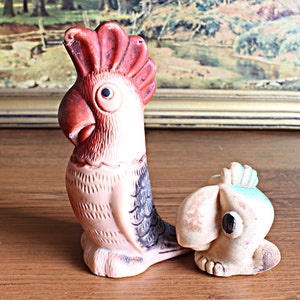 May include: A vintage, hand-painted parrot figurine with a red crest, brown beak, and black eye. The bird is light pink with black and gray wing details. A smaller, matching bird figurine is also present.