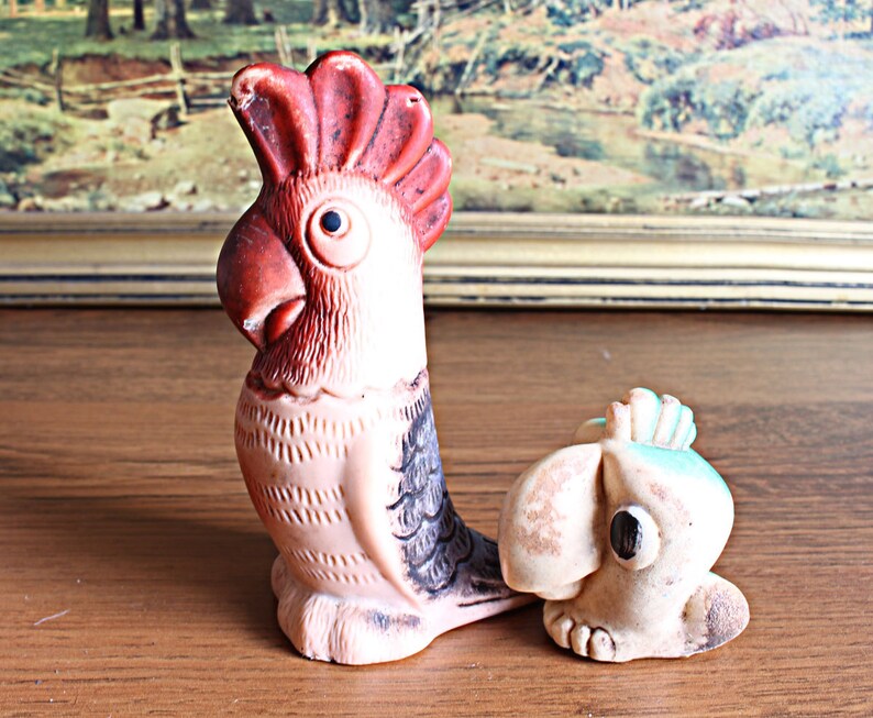 May include: A vintage, hand-painted parrot figurine with a red crest, brown beak, and black eye. The bird is light pink with black and gray wing details. A smaller, matching bird figurine is also present.