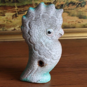 May include: A ceramic bird figurine in shades of gray and turquoise. The bird has a textured surface and a hole on its side. The figurine is perched on a wooden surface.