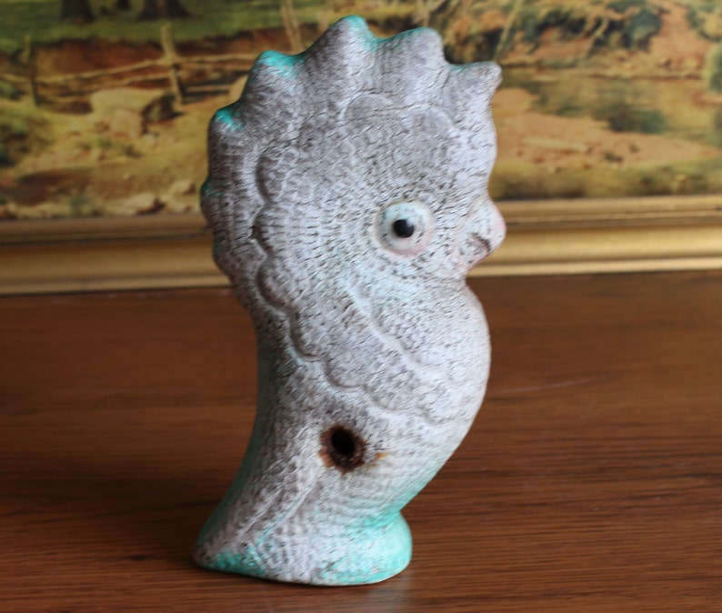 May include: A ceramic bird figurine in shades of gray and turquoise. The bird has a textured surface and a hole on its side. The figurine is perched on a wooden surface.