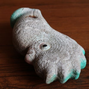May include: A weathered, abstract sculpture in the shape of a bird's foot. The sculpture is primarily gray with turquoise accents on the toes and head. The surface has a cracked, aged appearance, suggesting it is made of stone or a similar material.