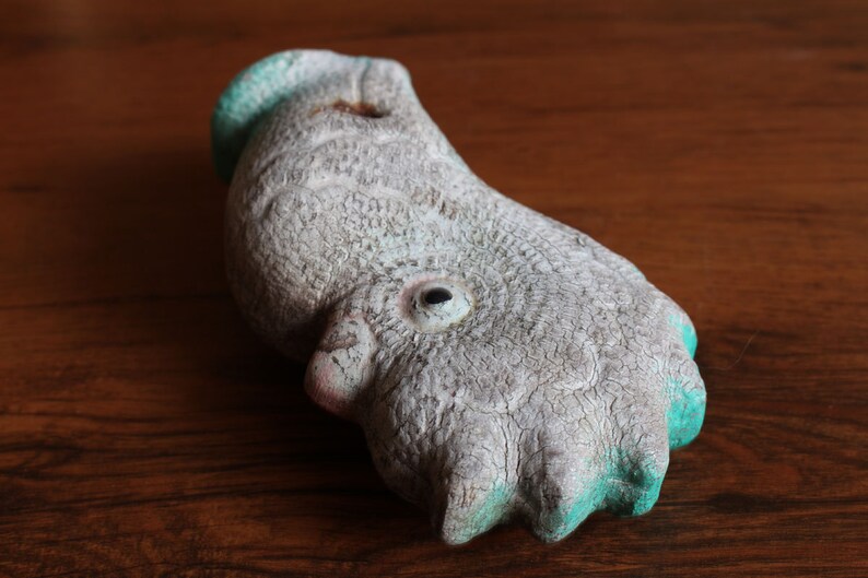 May include: A weathered, abstract sculpture in the shape of a bird's foot. The sculpture is primarily gray with turquoise accents on the toes and head. The surface has a cracked, aged appearance, suggesting it is made of stone or a similar material.