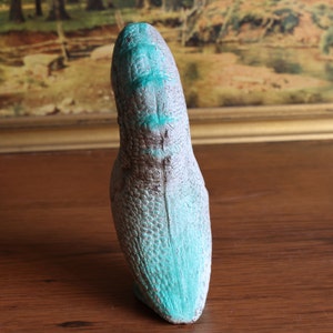 May include: A gray and brown dinosaur head sculpture with turquoise accents. The sculpture has a textured, scaled surface and is displayed on a wooden surface. The artwork is a unique decorative piece.