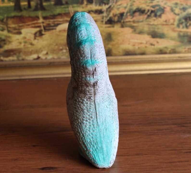 May include: A gray and brown dinosaur head sculpture with turquoise accents. The sculpture has a textured, scaled surface and is displayed on a wooden surface. The artwork is a unique decorative piece.