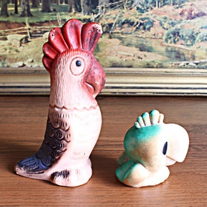 May include: Two vintage parrot figurines. One is a tall, upright parrot with a red crest, pink body, and dark blue accents. The other is a smaller, squat parrot in shades of green, blue, and cream. Both are on a wooden surface.
