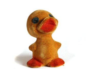 May include: A vintage, plush duck toy with a golden-brown body, orange feet and beak, and blue eyes. The duck is sitting upright, with its head tilted slightly upwards. The toy has a soft, velvety texture.