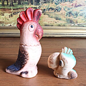 May include: Two vintage parrot figurines. One is tall and upright, with a red crest, pink body, and blue and black accents. The other is smaller, with a blue crest and a tan body. Both have a distressed, aged appearance.