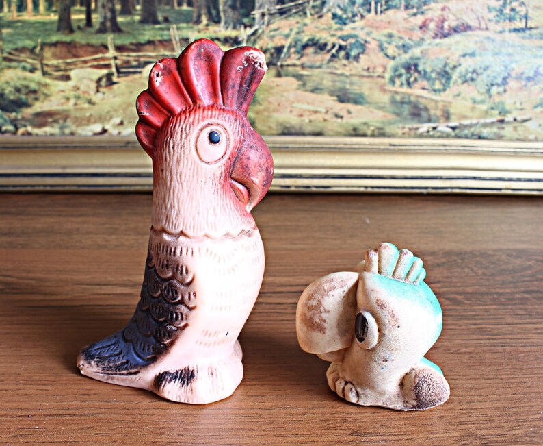 May include: Two vintage parrot figurines. One is tall and upright, with a red crest, pink body, and blue and black accents. The other is smaller, with a blue crest and a tan body. Both have a distressed, aged appearance.