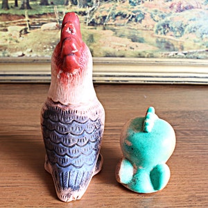 May include: Two ceramic bird figurines. One is a tall rooster with a red head and a cream-colored body with blue and black detailing. The other is a small, turquoise bird with a spiky crest.