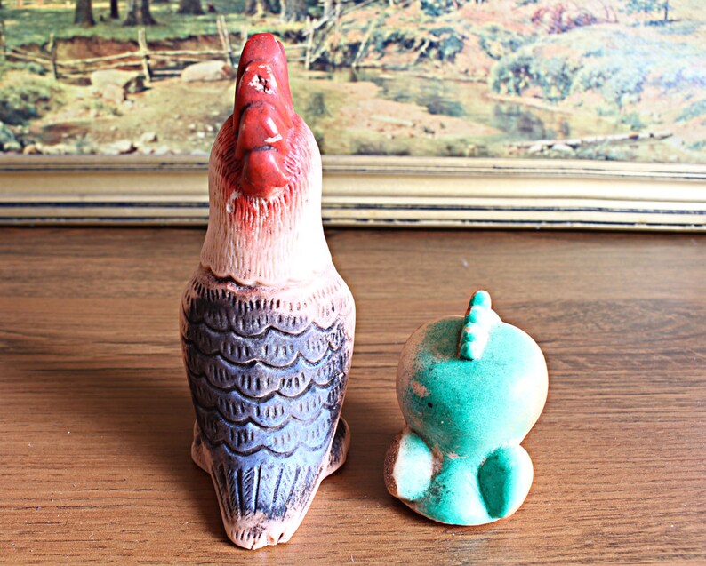 May include: Two ceramic bird figurines. One is a tall rooster with a red head and a cream-colored body with blue and black detailing. The other is a small, turquoise bird with a spiky crest.