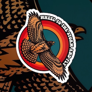 May include: A sticker featuring a stylized eagle in flight, with orange and brown feathers, set against a rainbow-colored circle. The text "WHEEL TO THE STORM AND FLY" arcs around the circle. The background has a repeating pattern of eagle wings.