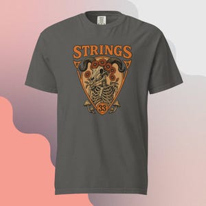 May include: Dark gray t-shirt with the word "STRINGS" in orange block letters above a graphic. The graphic features a skull with horns, flowers, and a skeleton torso inside an orange triangle. The number "33" is below the graphic.