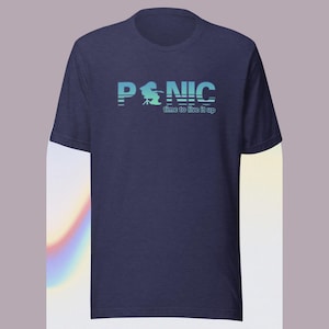 May include: Navy blue t-shirt with the word "PANIC" in a stylized, gradient blue font. Below the word is the phrase "time to live it up" in a smaller font. The shirt has short sleeves and a crew neck.