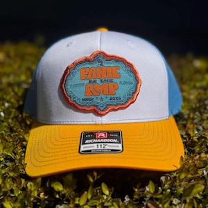 May include: A trucker hat with a white front panel, blue mesh back, and a yellow brim. The hat features a patch with the words "Panic at the RMP" in orange and blue, with "Florida" and "March 2025" text.