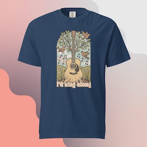 May include: Navy blue t-shirt featuring a guitar design with birds, musical notes, and the text "I'd sing along." The graphic is in shades of brown, beige, and green, creating a nature-inspired theme. The shirt is made of a soft, comfortable material.